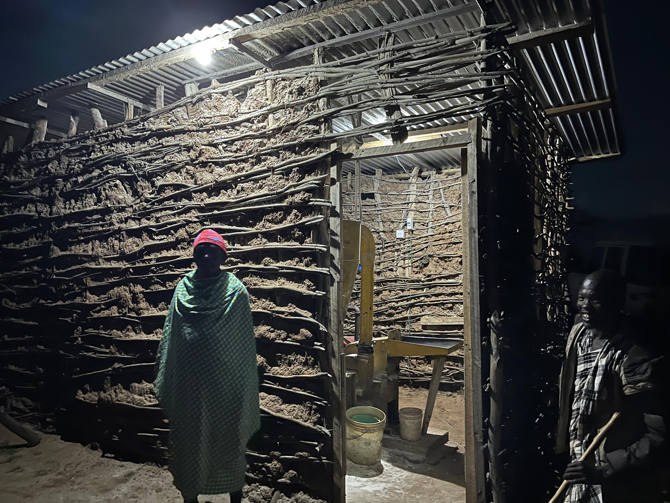 A person stands inside a small mud hut with his milling machine, illuminated by a clean electric light bulb.